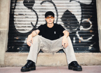 Downtown Sai in a cap and sunglasses, sitting in front of a graffiti-covered shutter
