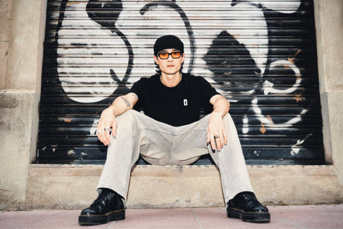 Downtown Sai in a cap and sunglasses, sitting in front of a graffiti-covered shutter